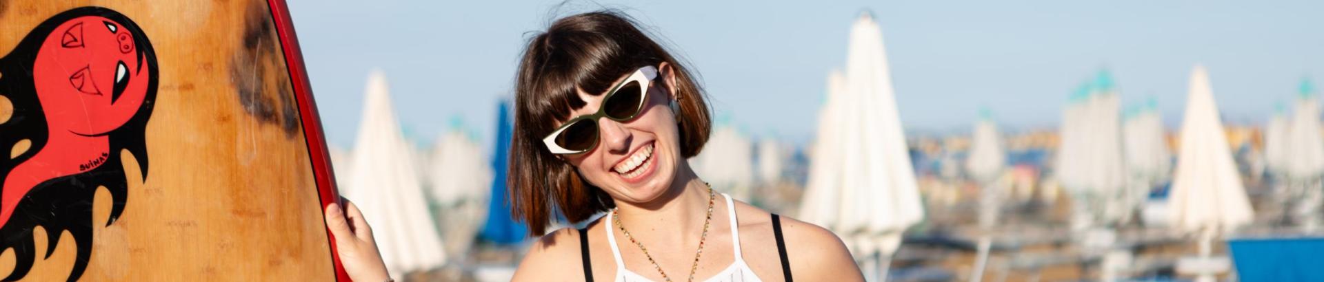 Smiling woman with sunglasses, at the beach with a surfboard.